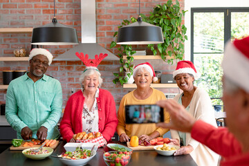 Group of happy diverse senior male and female friends in christmas hats taking selfie in kitchen