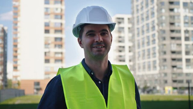 Man wearing hard hat and safety vest smiling at moving camera, parallax effect. Portrait of civil engineer or building contractor against multistory houses. Concept of construction industry