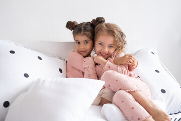 two beautiful diverse kids girls in pajamas hugging on bed in modern bright apartment
