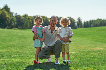 Caring dad smiling at camera, posing together with his cute little daughter and son, standing together on grass field in summer park