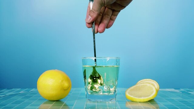 Stirring fresh lemon juice with ice in glass on table with blue background, bartender prepare fruit cocktail or fresh juice for drinking in party at bar