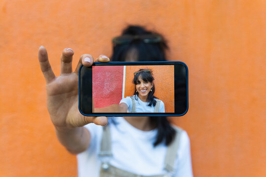 Young woman showing photograph on smart phone in front of orange wall
