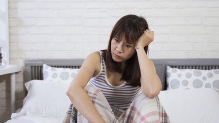 young asian chinese woman sitting with her arms on knees, looking outside and downward, doesn't want to go to work in the morning on the white bed