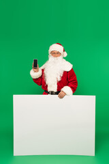Santa claus holding smartphone with blank screen and placard on green background