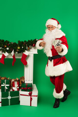 Santa claus pointing at dollars near fireplace and gifts on green background