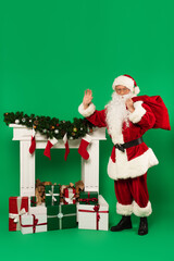 Santa claus with sack waving at camera near fireplace and presents on green background