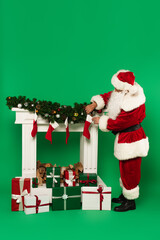 Santa claus holding christmas stocking near decorated fireplace and presents on green background