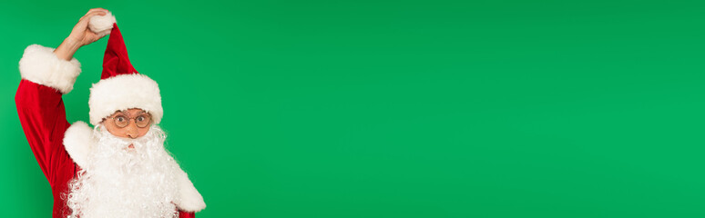 Santa claus holding hat and looking at camera isolated on green, banner