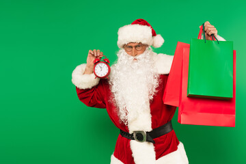 Santa claus in costume holding alarm clock and shopping bags isolated on green