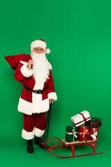 Santa claus with sack standing near sleigh with gifts on green background
