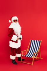 Santa claus in costume and sunglasses standing near deck chair on red background