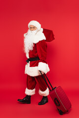 Santa claus in costume holding suitcase and sack on red background