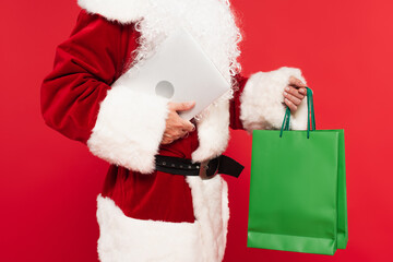 Cropped view of santa claus holding shopping bags and laptop isolated on red