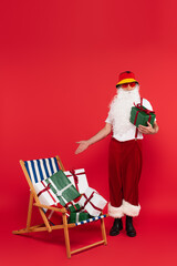 Santa claus in sunglasses and panama pointing at presents on deck chair on red background