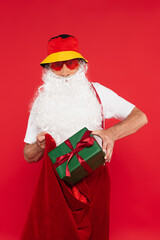 Santa claus in panama and sunglasses putting present with bow in sack isolated on red