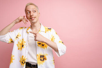 Young blonde man pointing finger aside and making handset gesture