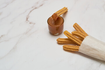 Traditional churros with sugar and cinnamon served on a marble table, with a bowl of caramel.