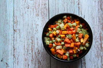 Vegetables, raw, cut very small, with carrots, green beans, pumpkin and chayote, in a black bowl on a wooden table, isolated.