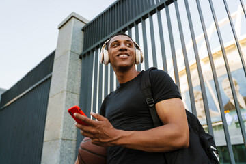 Black sportsman listening music while standing with basketball