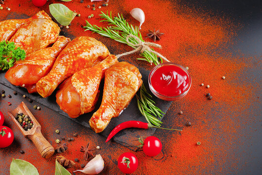 Barbecue marinated chicken dramstik on a black cooking board, with fresh rosemary leaves and fresh cherry tomatoes and various spices, close-up.Semi-finished product, Marinated product, quick cooking.