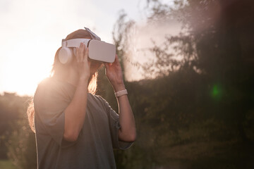 The girl, wearing virtual reality glasses, holds the glasses with her hands. Against the background of green trees. There is a beam of sunlight nearby. High quality photo