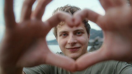 Young blond boy towards camera making heart symbol with hand and smiling looking at camera