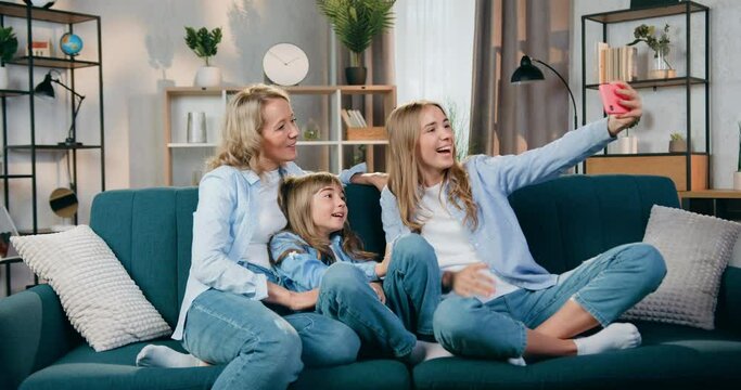 Attractive happy positive friendly mother and her two daughters spending leisure at home and having video call on mobile,they waving hands to say hallo to relatives