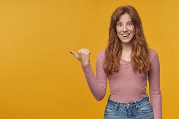 Indoor portrait of young ginger happy female with wavy hair and freckles indicates with a finger at copy space aside, smiles broadly. Isolated