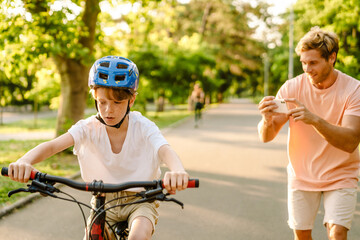 Naklejka premium Ginger white man taking photo on cellphone while his son riding bicycle