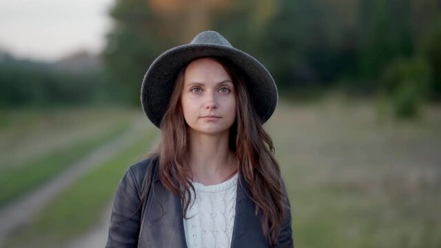 A long-haired young woman in a hat is walking alone in the park, looking sadly at the camera