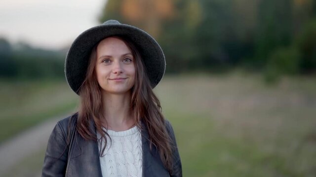 A long-haired young woman in a hat walks alone in the park, looks at the camera and smiles slightly