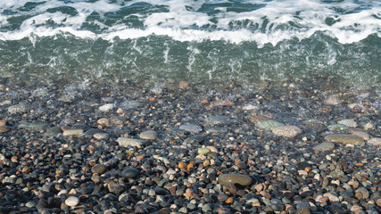 Sea surf on a sunny day. Light waves, pebble shore. Multi-colored wet pebbles on the shore