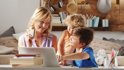 Happy family at home shopping online with credit card, mother with son and daughter have fun using computer, together sitting in living room at home, they surf internet looking for a purchase for gift