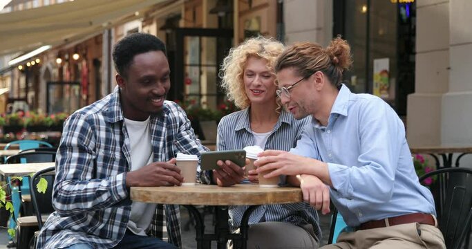 Handsome multiracial man sitting at the cafe with his friends and showing something on smartphone outdoor at street. People talking and using mobile in town