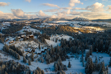 Aerial winter landscape with small village houses between snow covered forest in cold mountains in the evening.