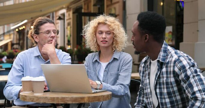 Mixed-races young people sitting at cafeteria terrace and working on laptop outdoor. Blonde girl showing something on computer to her friends. Leisure concept
