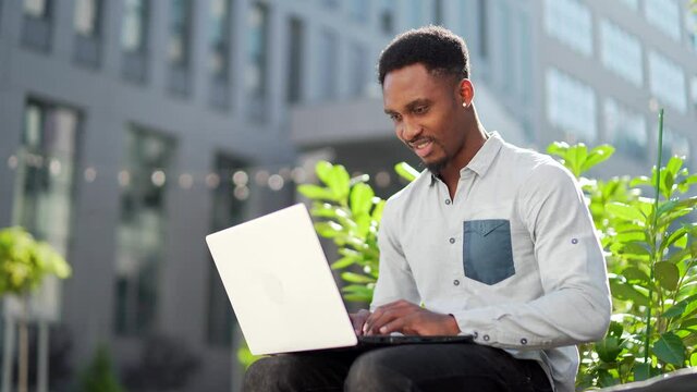 black male freelancer working online using laptop sitting on bench outside office modern building in city urban park on street. Happy African american guy student in casual outdoors typing on keyboard