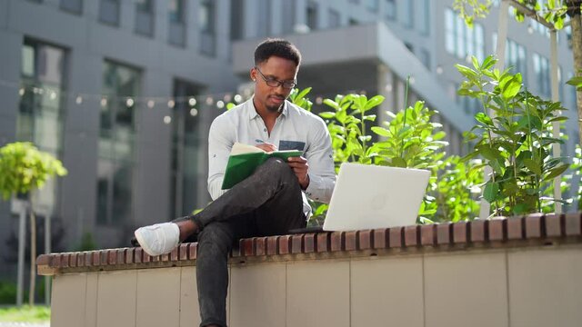Young african american male student sitting in a city park on a bench with laptop and notebook studying online outdoors Man freelancer in glasses with books learn working remotely in street e-learning