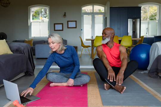 Happy senior diverse couple in exercise clothes practicing yoga together, using laptop