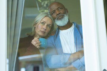 Worried senior diverse couple looking through window and embracing