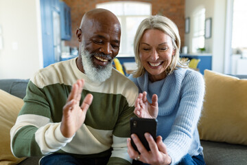 Happy senior diverse couple in living room sitting on sofa, using smartphone