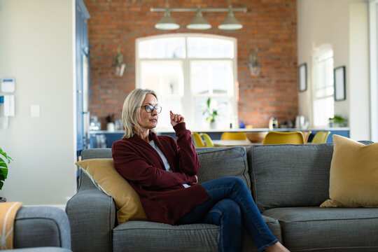 Thoughtful senior caucasian woman in living room sitting on sofa, thinking