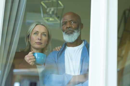 Worried senior diverse couple looking through window and embracing
