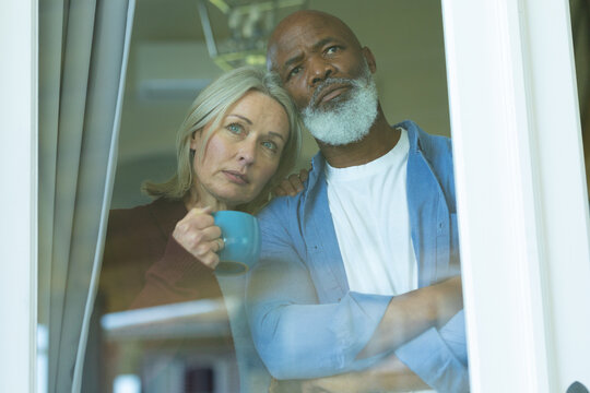 Worried senior diverse couple looking through window and embracing