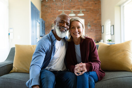 Portrait of happy senior diverse couple in living room sitting on sofa, embracing and smiling