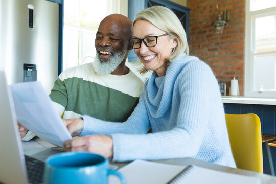 Happy senior diverse couple in kitchen sitting at table, using laptop - Powered by Adobe