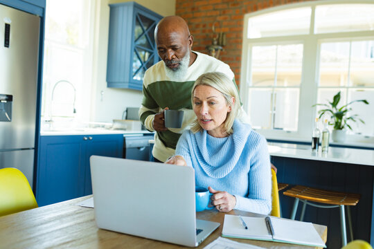Stressed senior diverse couple in kitchen sitting at table, using laptop