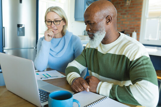 Stressed senior diverse couple in kitchen sitting at table, using laptop