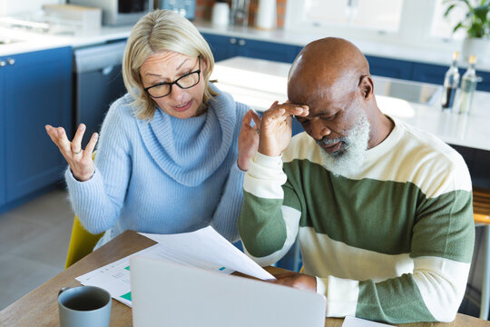 Stressed senior diverse couple in kitchen sitting at table, using laptop
