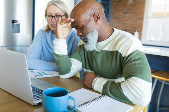 Stressed senior diverse couple in kitchen sitting at table, using laptop